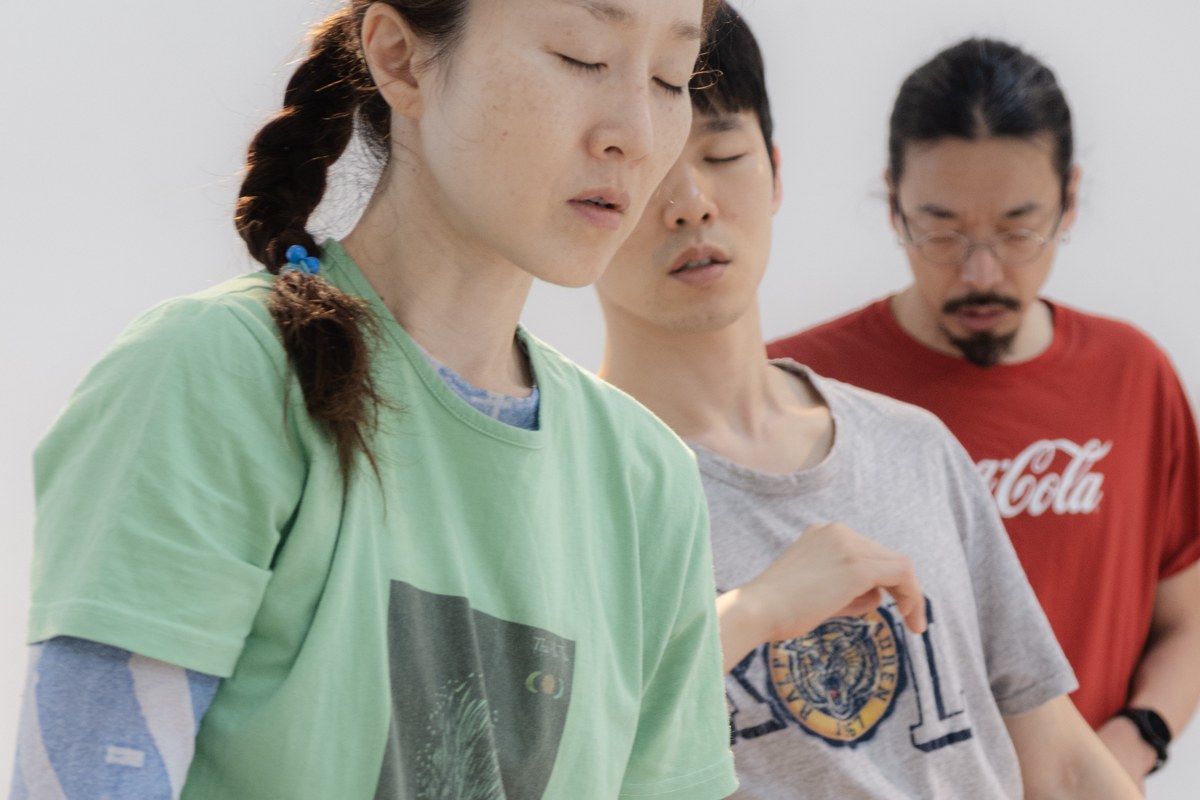 <건져올렸지만 가라앉으려는 경향>의 한 장면. 밝은 흰 벽 앞에 세 사람이 앞뒤로 겹쳐 서 있다. 앞쪽의 머리를 땋은 사람은 초록색 티셔츠를 입고 눈을 감은 채 고개를 살짝 숙이고 있다. 뒤쪽 두 사람도 눈을 감고 있으며, 맨 뒤 사람은 안경을 쓰고 붉은 티셔츠를 입었다.