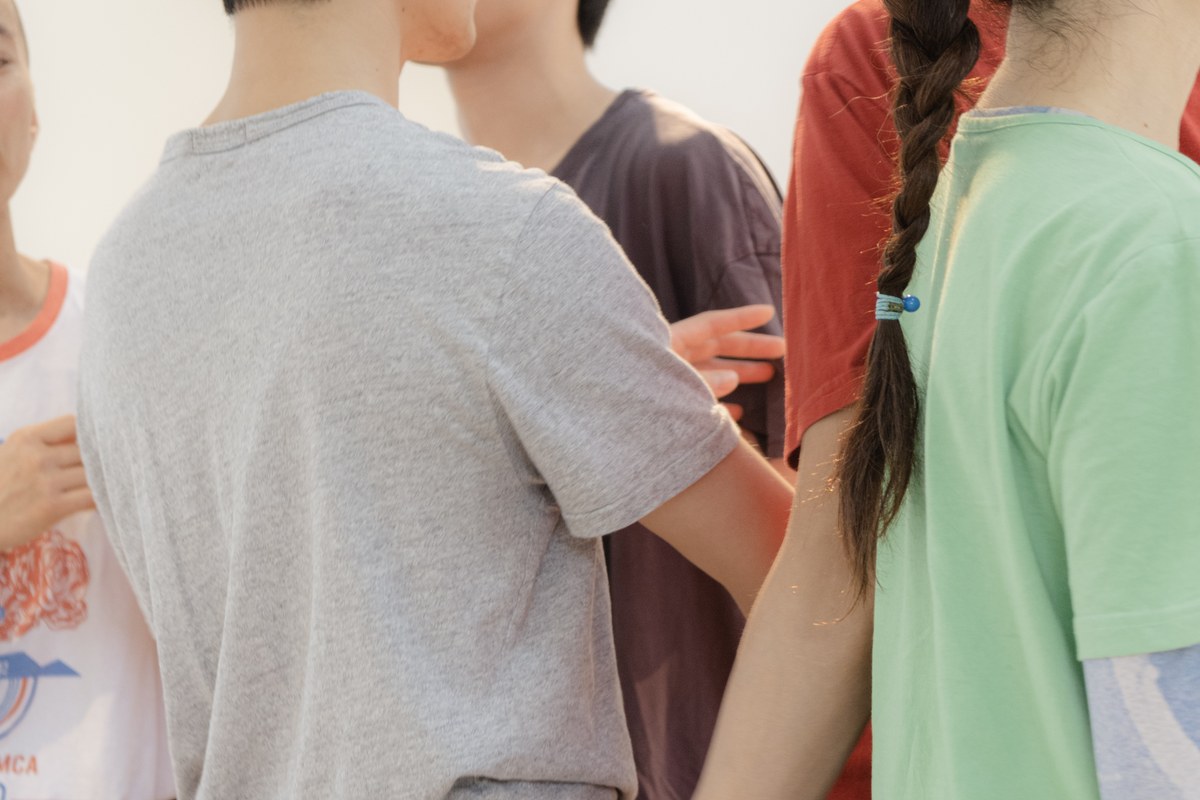 <건져올렸지만 가라앉으려는 경향>의 한 장면. 밝은 공간에서 여러 사람이 가까이 모여 서 있다. 화면에는 얼굴 대신 상체와 팔, 서로 닿은 손이 담겨 있다. 회색 티셔츠를 입은 사람의 등 뒤로 초록색 티셔츠를 입고 머리를 땋은 사람이 손을 뻗어 어깨에 대고 있다.