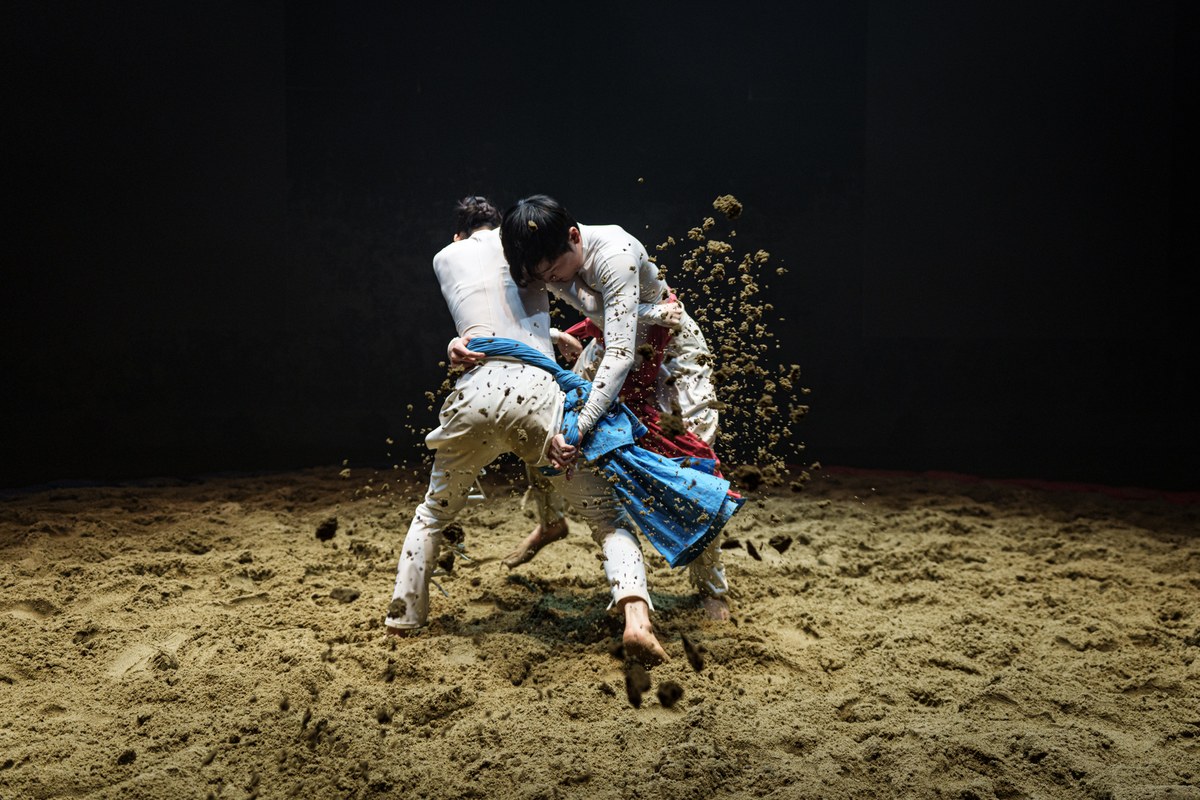 <씨름>의 한 장면. 모래판 위에서 흰옷을 입은 두 무용수가 서로 샅바를 잡고 힘껏 맞붙어 있다. 한 사람이 상대를 넘기려는 순간 모래가 공중으로 크게 흩날린다. 파란색과 붉은색 샅바가 두 사람 사이에서 뒤엉켜 있다.