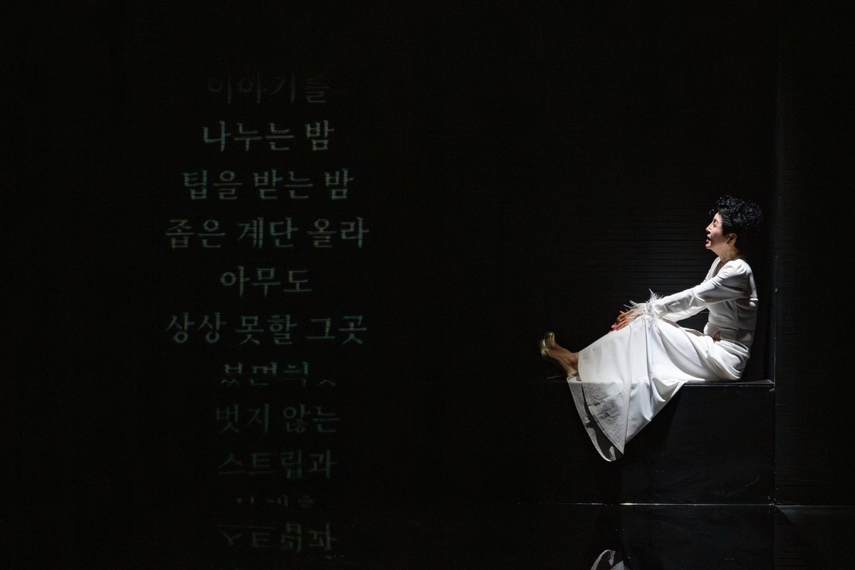 <“뺨을 맞지 않고 사는 게 삶의 전부가 될 순 없더라”>의 한 장면. 어두운 무대 왼쪽에 ‘이야기를 나누는 밤 / 팁을 받는 밤 / 좁은 계단 올라 / 아무도 상상 못할 그곳’이라는 자막이 세로로 떠 있다. 오른쪽 검은 단 위에는 흰옷을 입은 인물이 다리를 뻗고 앉아 있으며, 반들거리는 바닥에 그 모습이 거울처럼 비친다.
