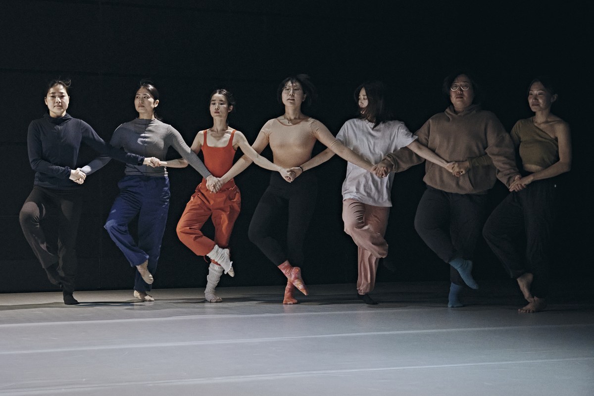 <메타발레: 비(非)-코펠리아 선언>의 한 장면. 검은 배경 앞에 일곱 명의 무용수가 옆으로 나란히 서서 서로 손을 맞잡고 있다. 모두 한쪽 다리를 들어 발끝을 반대쪽 무릎에 붙인 발레 자세를 취하고 있으며, 각자 다른 색과 스타일의 평상복을 입고 있다.