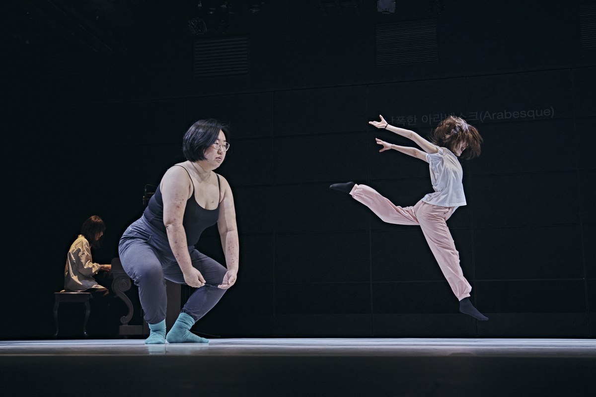 <메타발레: 비(非)-코펠리아 선언>의 한 장면. 어두운 무대 왼쪽에서 검은 민소매 상의를 입고 안경을 쓴 무용수가 무릎을 굽히고 몸을 낮추고 있다. 오른쪽에서는 분홍색 바지를 입은 무용수가 한쪽 다리를 뒤로 뻗고 두 팔을 앞으로 펼치며 공중으로 뛰어오른다. 뒤쪽 벽에는 ‘아라베스크(Arabesque)’라는 글자가 희미하게 비치고, 왼쪽 뒤에서는 한 사람이 피아노를 연주하고 있다.
