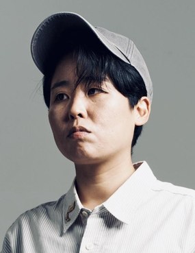 구자혜 연출가의 프로필 사진. 회색 배경 앞에서 회색 모자를 쓰고 흰 줄무늬 셔츠를 입은 사람이 정면을 바라보고 있다.