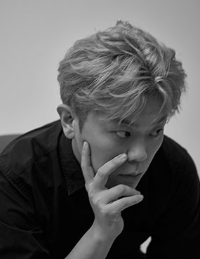 음악가이자 연출가인 김재훈의 프로필 사진. 밝은 색으로 염색한 짧은 머리를 자연스럽게 넘기고 검은색 긴팔 셔츠를 입었다. 고개를 약간 숙인 채 한 손으로 턱과 뺨을 받치며 오른쪽을 본다. 흑백사진이다.