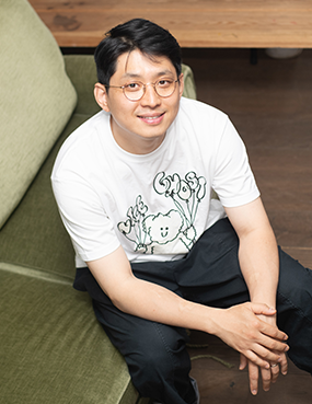 연극 예술가인 구자하의 프로필 사진. 짧은 검은 머리를 옆으로 넘기고 둥근 안경을 썼다. 곰돌이가 풍선을 들고 있는 그림이 그려진 흰 반팔 티셔츠와 검정 바지를 입었다. 카키색 소파에 앉아 팔을 허벅지에 올린 채, 미소를 지으며 정면을 바라본다.