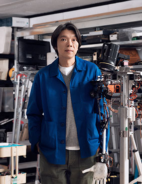 뉴미디어 퍼포먼스 기획자이자 연출가인 권병준의 프로필. 귀까지 오는 검은 머리를 옆으로 넘기고, 흰색 티셔츠와 회색 니트, 파란색 재킷과 카키색 바지를 입었다. 머리 대신 손전등이 달린 로봇의 팔 옆에 서서 정면을 바라보고 있다.